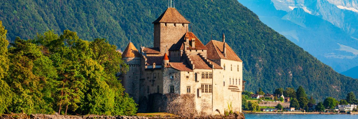 Historic Chillon Castle on the shores of Lake Geneva in Switzerland, surrounded by lush green mountains and alpine peaks under a clear blue sky.