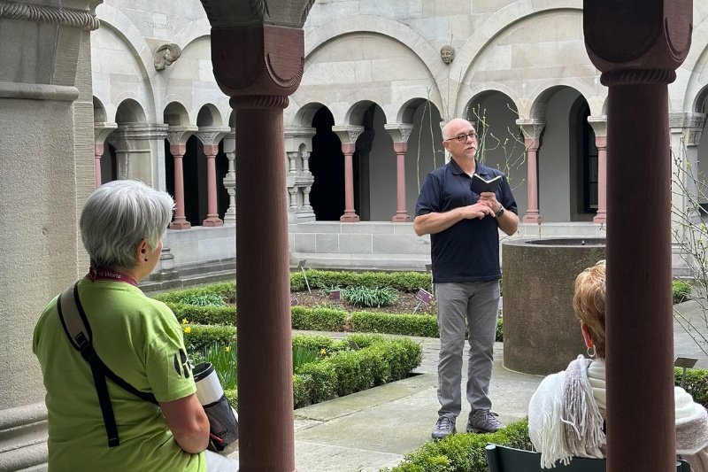 Rick Brumback speaking to visitors inside a historic garden with stone arches and greenery in Switzerland.