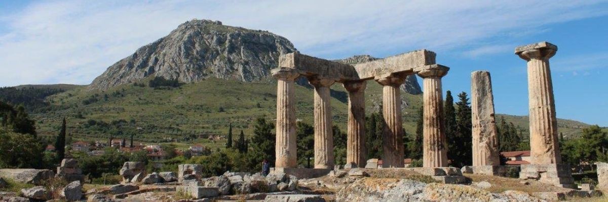 Ruins of the ancient Temple of Apollo in Corinth, Greece, with standing Doric columns and a scenic mountain backdrop under a bright, clear sky.