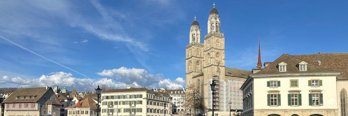 Scenic view of Zurich’s Old Town featuring the iconic Grossmünster Church twin towers, historic buildings, and people walking across a bridge under a bright blue sky.