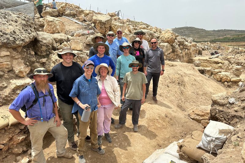 A large group of excavation team members posing on a dig site at Shiloh, wearing hats and gear, with stone ruins and hills in the background.