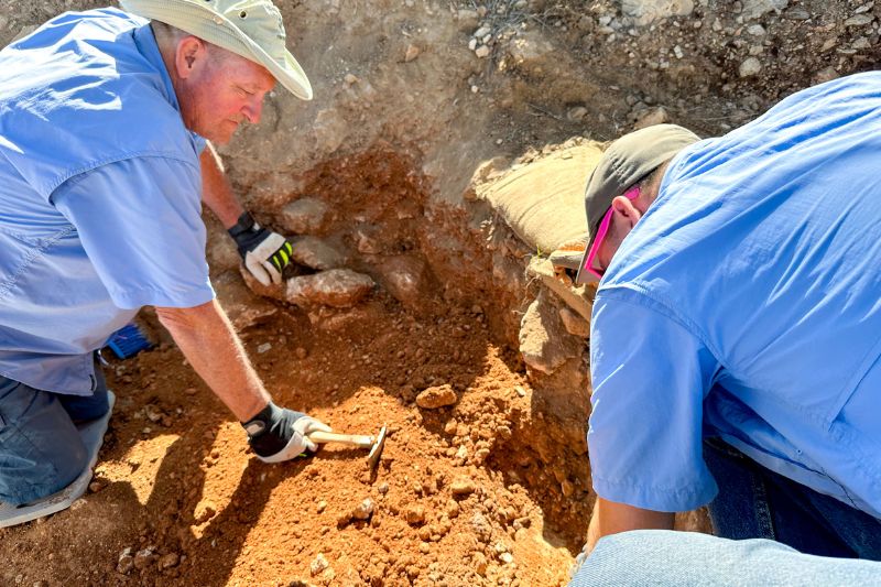 Two archaeologists carefully excavating soil with hand tools in a trench at Shiloh, examining the reddish earth for artifacts.