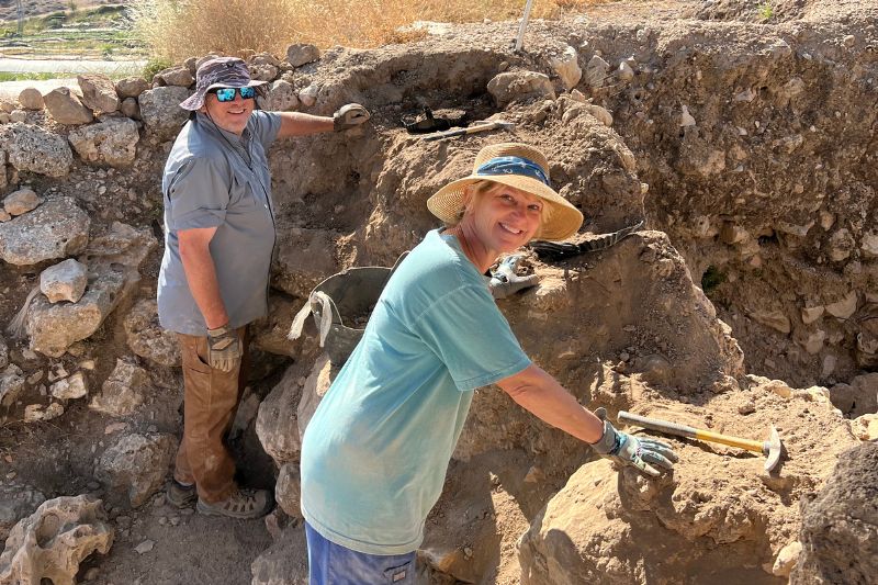 Two archaeologists smiling while working in an excavation site at Shiloh. They are digging and brushing dirt from ancient stone walls under the sun.