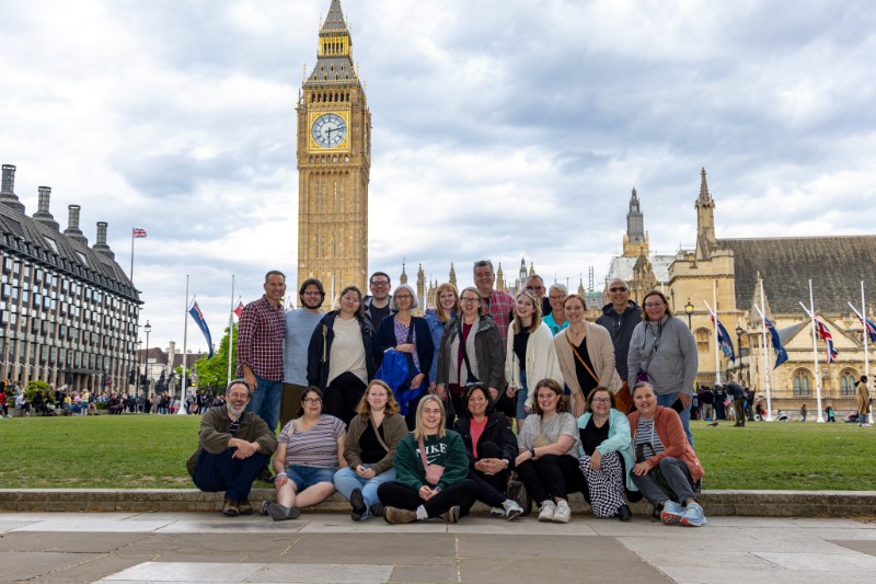 A large group of smiling travelers poses on the lawn in front of the iconic Big Ben clock tower and the Houses of Parliament in London, with flags and historic architecture visible in the background under a partly cloudy sky.