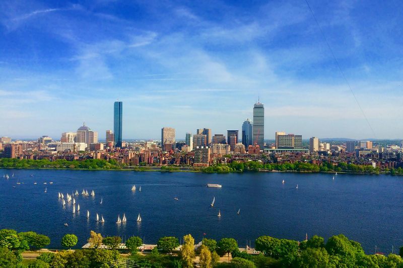 A panoramic view of the Boston skyline with sailboats dotting the Charles River on a clear day, surrounded by lush greenery and blue skies.