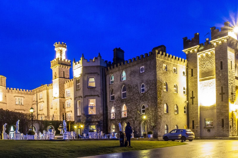 The illuminated stone facade of Cabra Castle stands against a deep blue evening sky, with statues, castle towers, and visitors gathered outside.