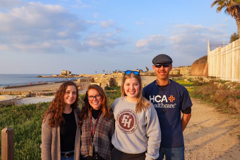 Four smiling travelers pose near the Mediterranean Sea at Caesarea Maritima, with ancient Roman ruins and palm trees under a partly cloudy sky.