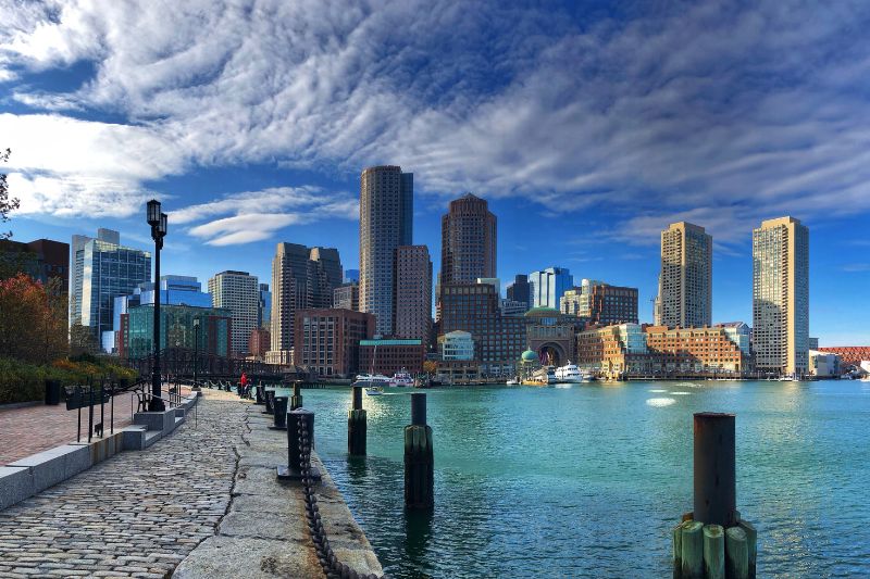 The Boston skyline as seen from the harbor, featuring tall modern skyscrapers, brick buildings, and a waterfront promenade under dramatic cloud cover.