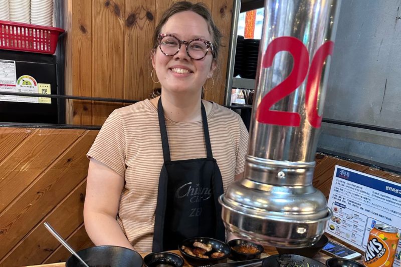 Woman smiling at a table set for Korean BBQ, with fresh meat grilling and traditional side dishes surrounding the hot plate.