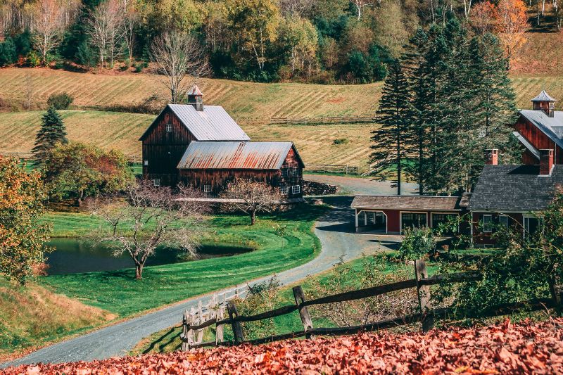 A scenic view of a rustic barn and farmhouse in Vermont surrounded by fall foliage, green pastures, and a winding gravel road.