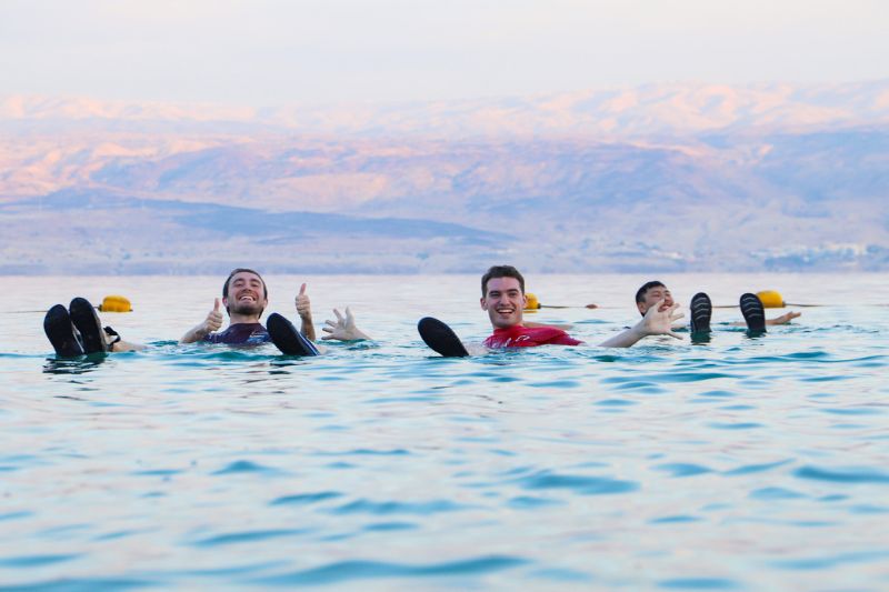 Young men floating effortlessly in the Dead Sea with smiles and thumbs up, surrounded by calm waters and the scenic backdrop of Jordan’s mountains.