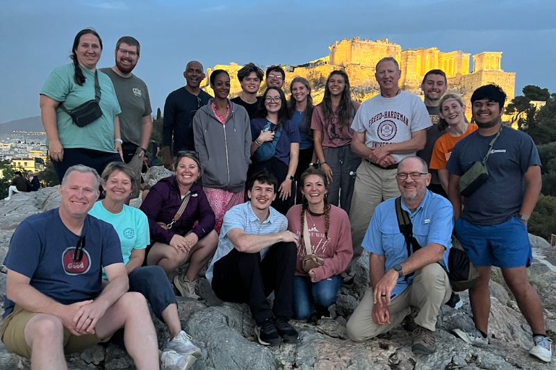 A group of travelers poses in front of the illuminated Acropolis in Athens, Greece at sunset during a Bible lands tour.