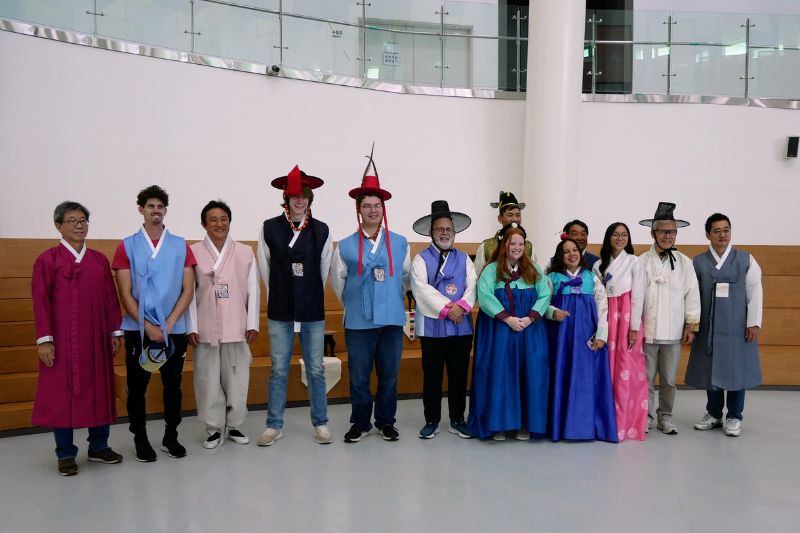 Group of people dressed in traditional Korean hanbok, smiling inside a modern building during a cultural exchange experience.