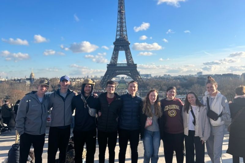 Group of students smiling in front of the Eiffel Tower on a clear day in Paris, France, with blue skies and city views in the background.