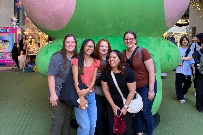 Group of young travelers smiling in front of a giant green plush frog character at a shopping center in South Korea, surrounded by vibrant lights and shops.
