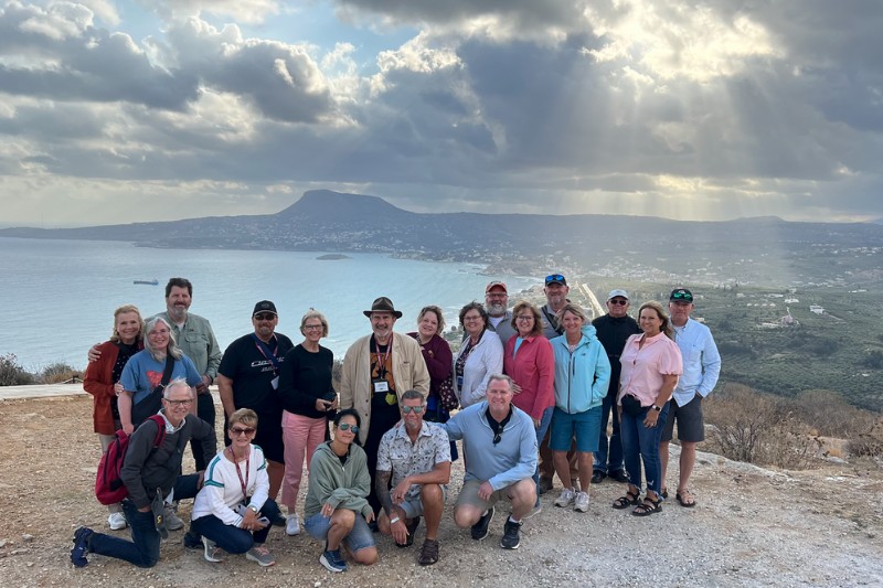 Travel group posing at a high-elevation lookout with a sweeping coastal landscape and dramatic sky in the background.