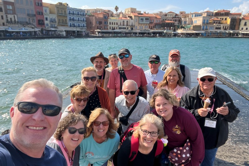 Group of tourists smiling at a scenic European harbor with colorful waterfront buildings in the background.