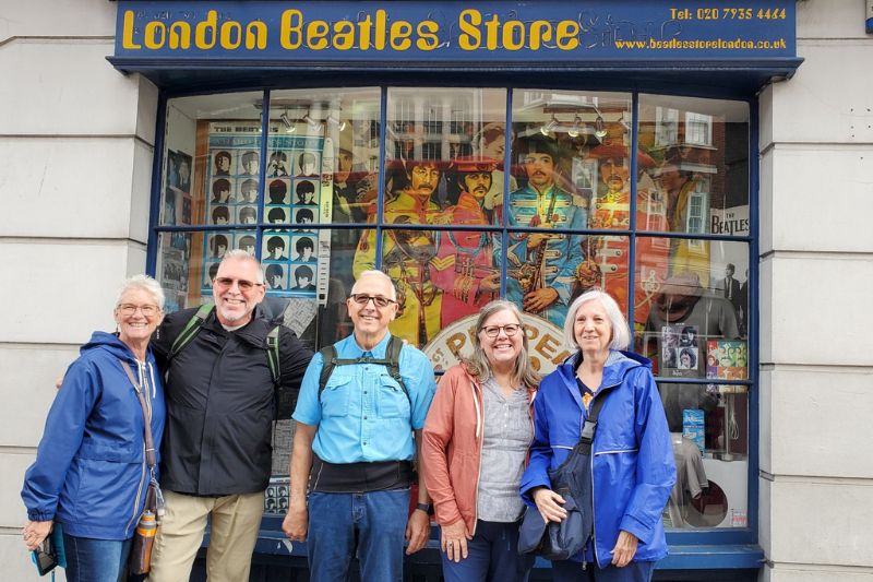 Five smiling adults stand in front of the London Beatles Store, with colorful Beatles-themed artwork and memorabilia visible through the shop window behind them.