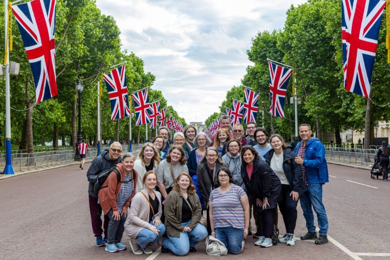 A large group of people poses for a photo in the middle of The Mall in London, with rows of Union Jack flags lining the street and lush trees framing the scene on both sides.