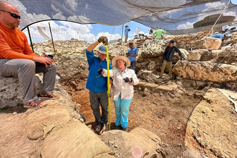 Two archaeologists measuring a trench depth with a tape measure at the Shiloh dig site, working under a mesh canopy for shade.