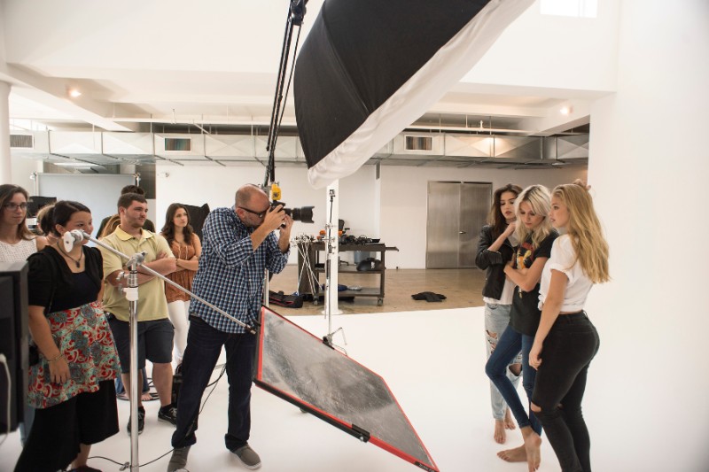 Photographer capturing three female models in a professional studio with lighting setup and crew observing.