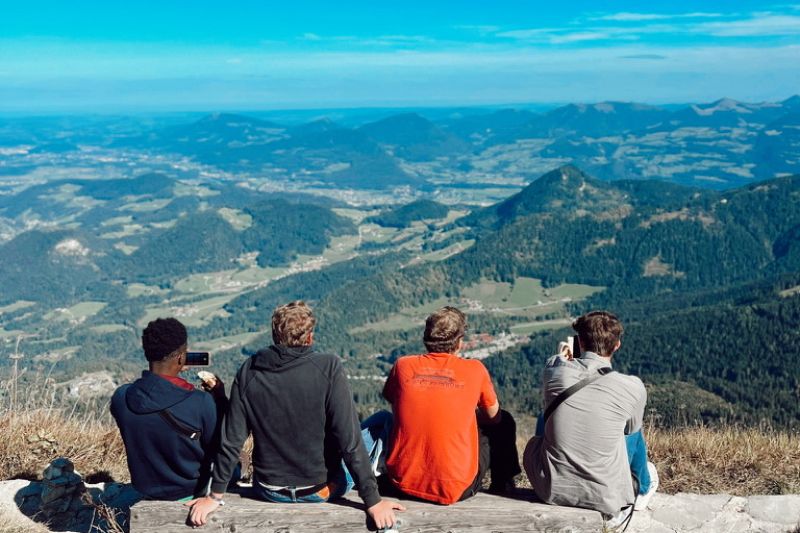 Four friends sitting on a scenic mountain overlook, capturing photos and enjoying panoramic views of rolling green hills and villages below in the Alps.