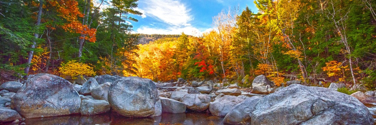 A vibrant autumn scene in New England featuring a rocky creek surrounded by colorful trees with golden, orange, and red foliage under a bright blue sky