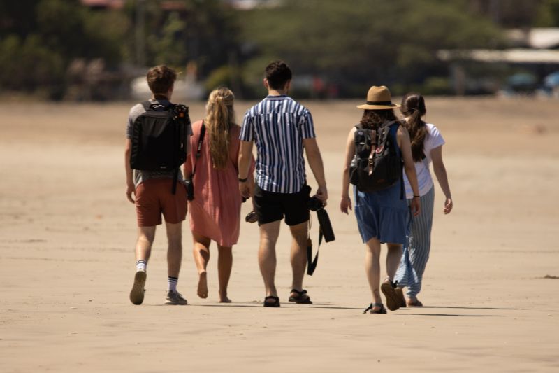 Group of friends walking barefoot on a sandy beach toward the horizon.