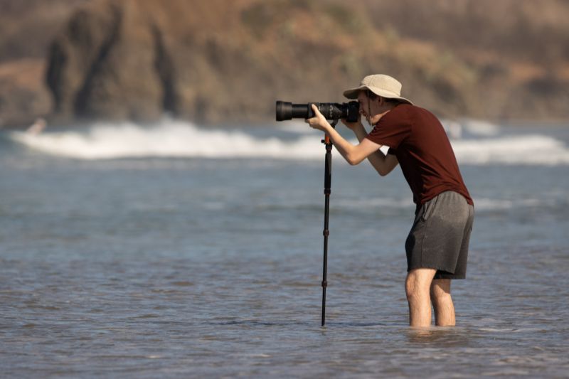 Photographer with a hat taking pictures in shallow ocean water using a monopod.
