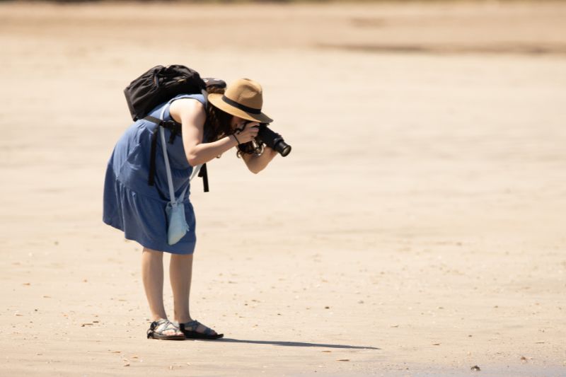 Woman in a blue dress taking beach photos with a DSLR camera and backpack.