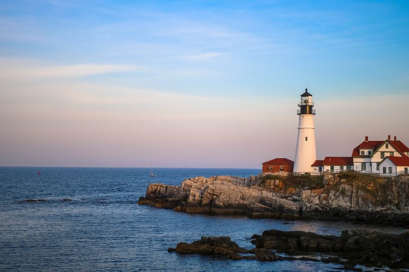 A historic white lighthouse with red-roofed buildings perched on rocky cliffs overlooking the Atlantic Ocean at sunset in Cape Elizabeth, Maine.