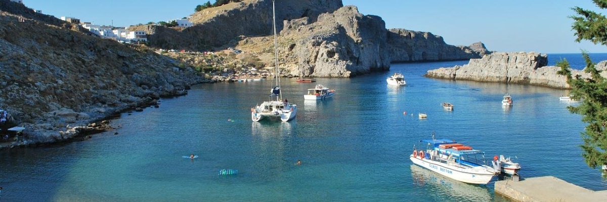 Clear blue bay with sailboats, swimmers, and a historic town built into rocky cliffs under a sunny sky.