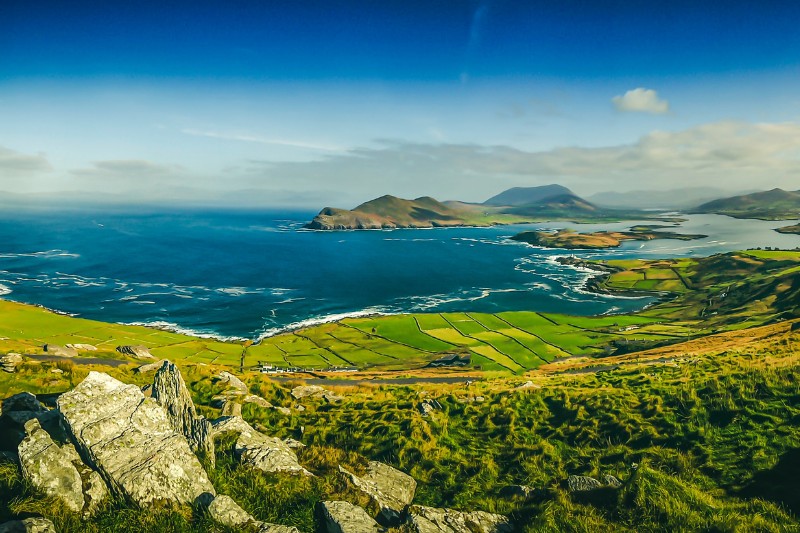A sweeping vista of Ireland's coastline from the Ring of Kerry, with rolling green fields, rocky cliffs, and deep blue waters stretching into the horizon.