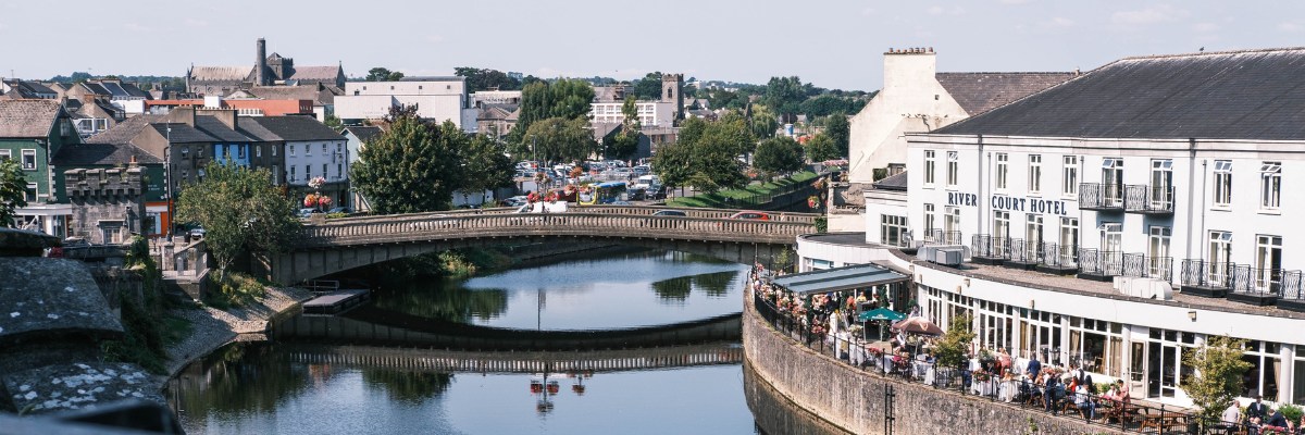 A peaceful river scene in Kilkenny, Ireland, featuring the River Court Hotel with outdoor diners along the curved stone embankment, a gently arched bridge, and a backdrop of charming town buildings under a clear sky.