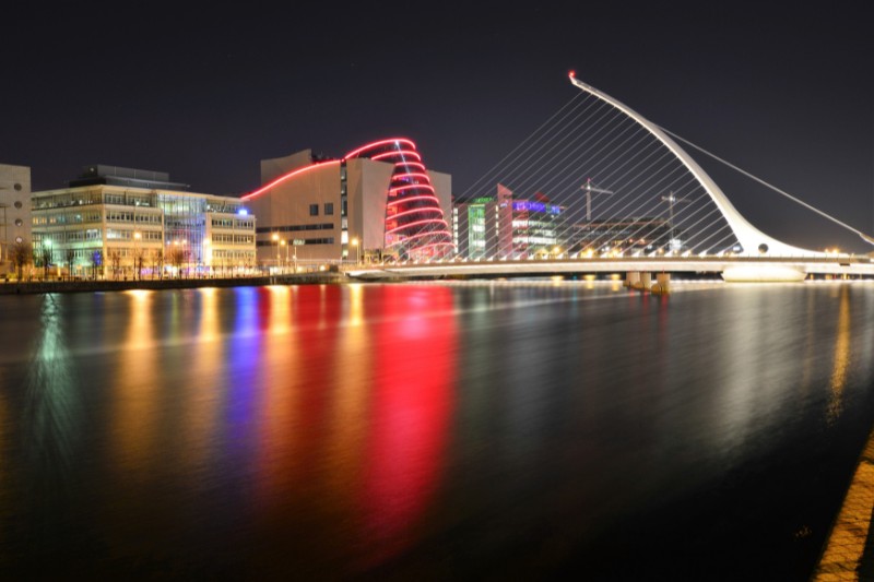 The Samuel Beckett Bridge in Dublin illuminated at night, with vibrant reflections of colorful lights on the River Liffey and the Dublin Convention Centre glowing red in the background.