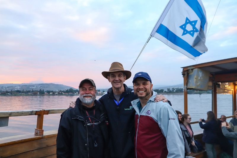Three men smiling on a wooden boat ride across the Sea of Galilee at sunset, with the Israeli flag waving in the breeze and mountains in the background.