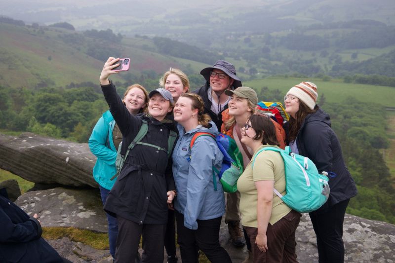 A group of young women dressed in rain jackets and backpacks smile and pose for a selfie at the top of a scenic overlook at Stanage Edge, surrounded by misty green hills and countryside.