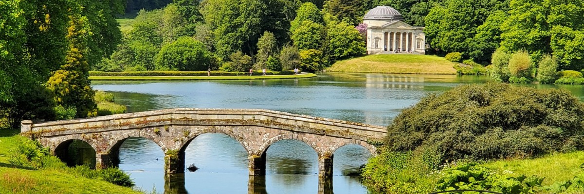 A scenic view of a stone footbridge crossing a tranquil lake, surrounded by lush greenery. In the background, a neoclassical temple with columns and a domed roof stands atop a gentle hill, framed by dense trees under a bright blue sky.