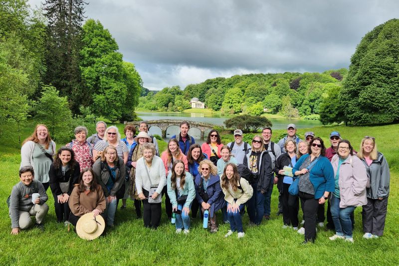 A cheerful tour group poses on the lawn at Stourhead Gardens, with a serene lake, arched stone bridge, and a classical temple in the background, surrounded by dense greenery and under a partly cloudy sky.