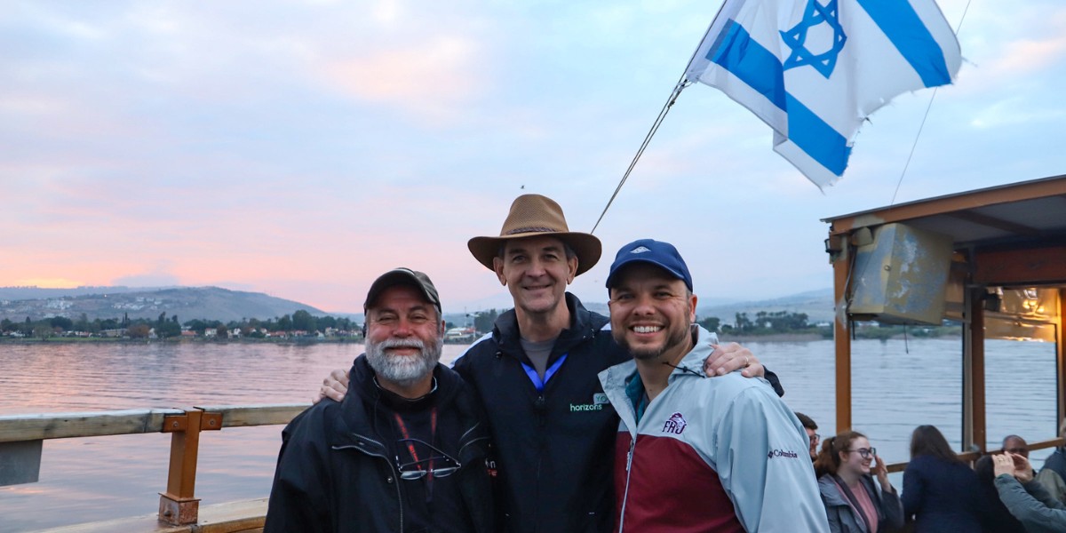 http://Three%20men%20smiling%20on%20a%20boat%20with%20the%20Israeli%20flag%20and%20Sea%20of%20Galilee%20in%20the%20background%20at%20sunset